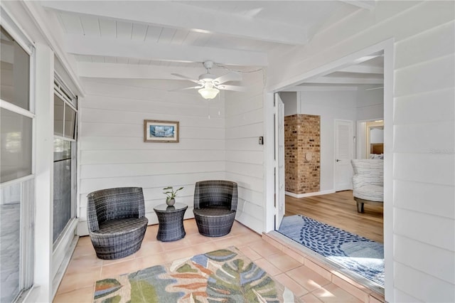 living area featuring a ceiling fan, a sunroom, wooden walls, beamed ceiling, and tile patterned floors
