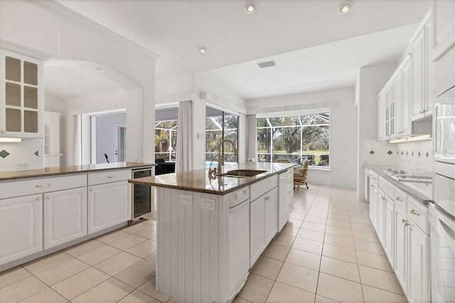 kitchen featuring light tile patterned floors, beverage cooler, a center island with sink, dark stone countertops, and glass insert cabinets