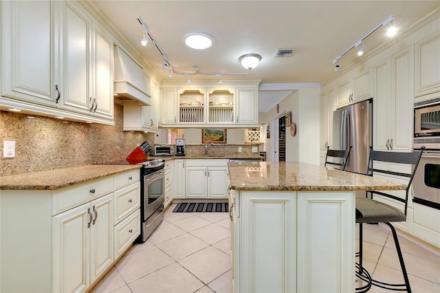 kitchen featuring light tile patterned floors, stainless steel appliances, custom range hood, glass insert cabinets, and light stone countertops