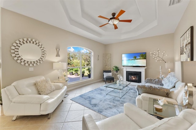 living room with a raised ceiling, visible vents, a ceiling fan, a glass covered fireplace, and light tile patterned flooring