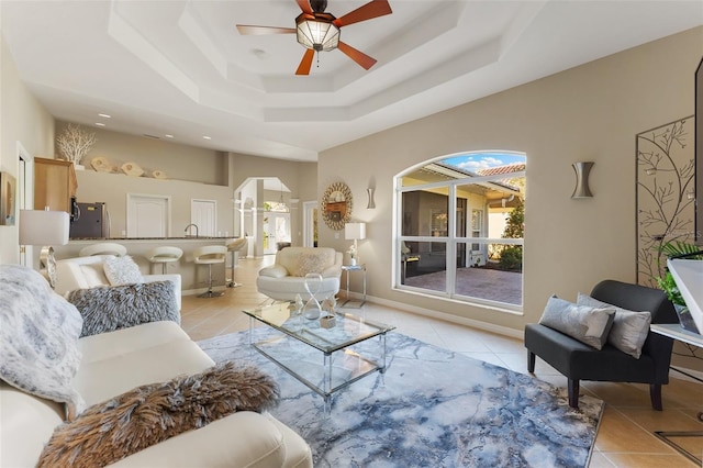 living area with light tile patterned floors, ceiling fan, a tray ceiling, and baseboards