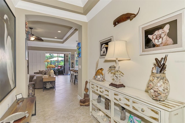 hall with a tray ceiling, crown molding, arched walkways, and light tile patterned floors