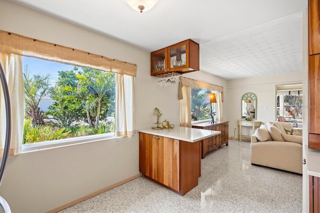 kitchen featuring light speckled floor, a peninsula, brown cabinetry, light countertops, and glass insert cabinets