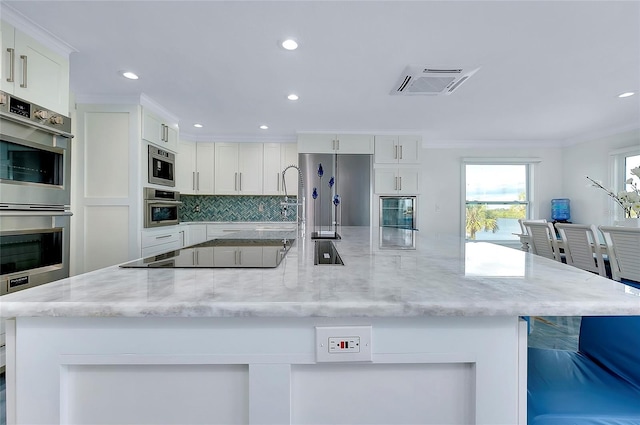 kitchen with tasteful backsplash, ornamental molding, stainless steel appliances, a large island with sink, and white cabinetry