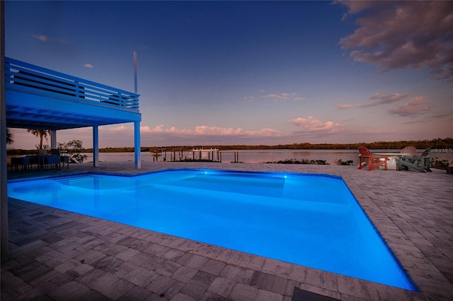 pool at dusk with a patio area, an outdoor pool, and a water view