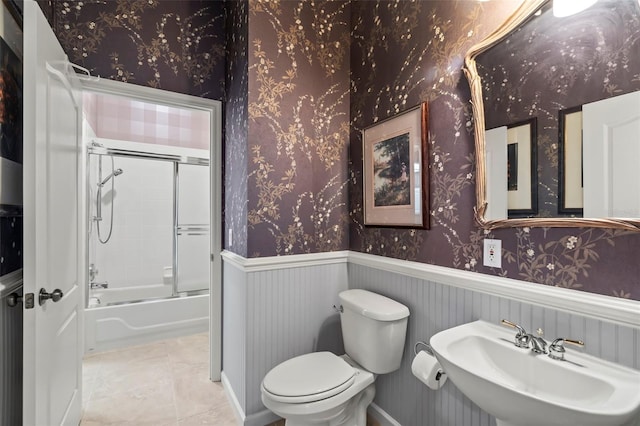 bathroom with wallpapered walls, toilet, shower / bath combination with glass door, a wainscoted wall, and a sink