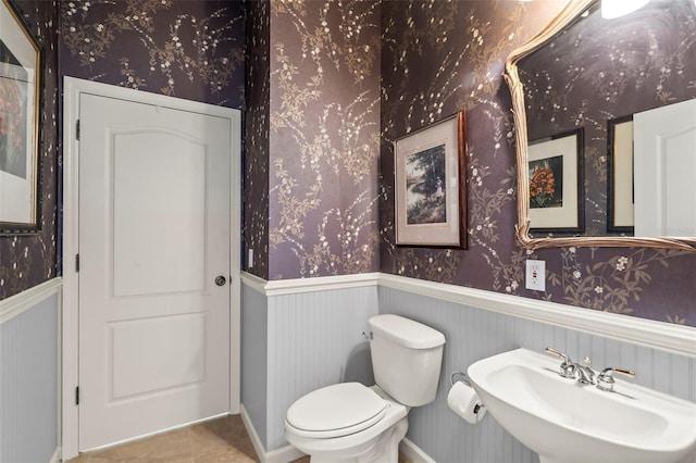 half bath with a wainscoted wall, toilet, a sink, tile patterned flooring, and wallpapered walls