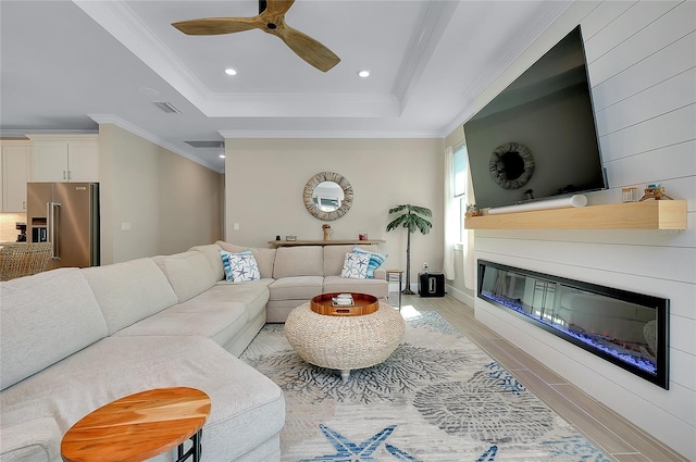 living area featuring a tray ceiling, a glass covered fireplace, ornamental molding, and light wood finished floors