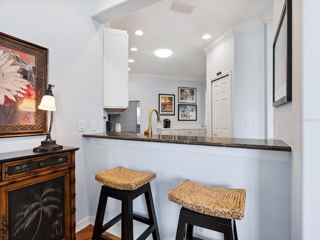 kitchen featuring a breakfast bar, white cabinetry, visible vents, dark stone countertops, and crown molding