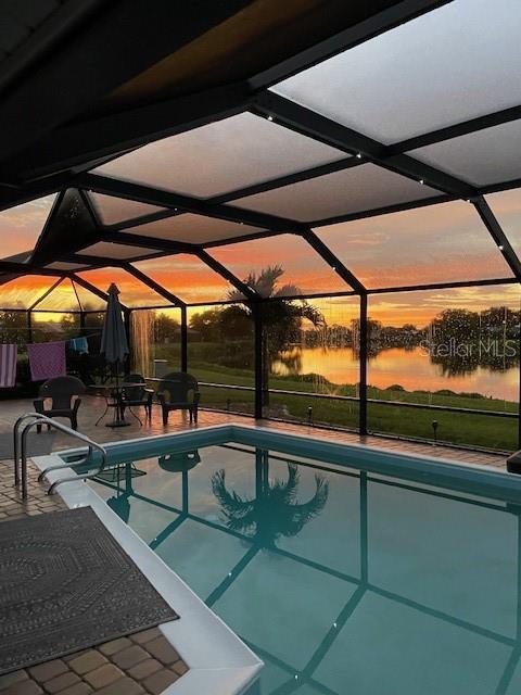 pool at dusk featuring a water view, glass enclosure, an outdoor pool, and a patio