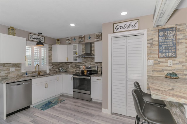 kitchen featuring appliances with stainless steel finishes, white cabinetry, a sink, wall chimney range hood, and light stone countertops