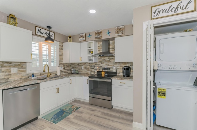 kitchen featuring stacked washer and dryer, a sink, white cabinetry, appliances with stainless steel finishes, and wall chimney exhaust hood