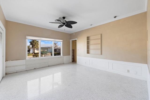 empty room with a ceiling fan, built in features, wainscoting, a decorative wall, and speckled floor