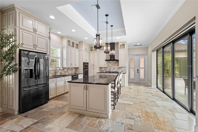 kitchen with stone tile flooring, a raised ceiling, black fridge with ice dispenser, a sink, and wall chimney exhaust hood