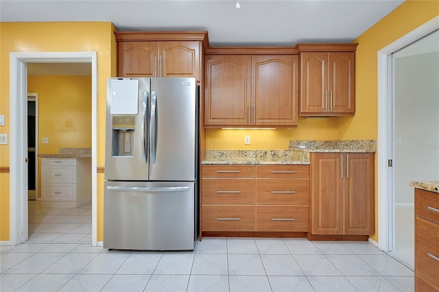 kitchen with brown cabinetry, baseboards, light stone countertops, stainless steel refrigerator with ice dispenser, and light tile patterned flooring