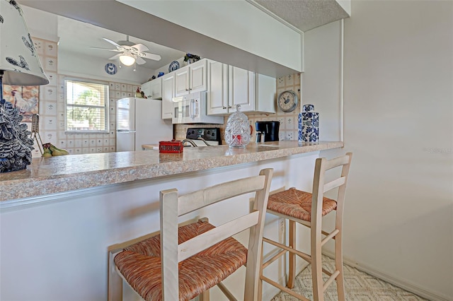 kitchen with white appliances, a ceiling fan, decorative backsplash, white cabinets, and a kitchen breakfast bar