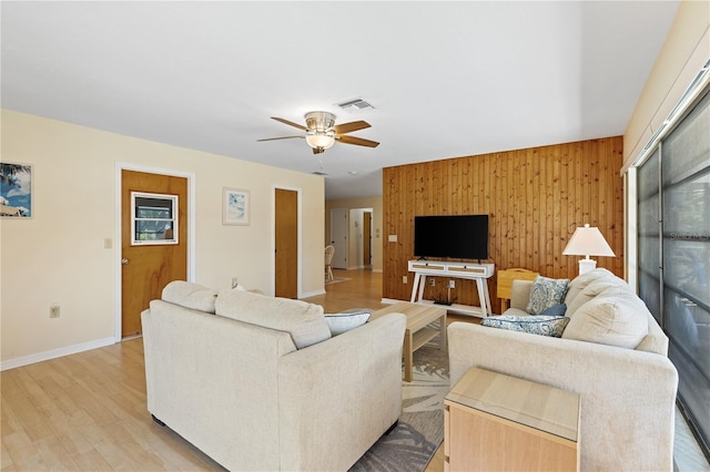 living room with baseboards, a ceiling fan, wood walls, visible vents, and light wood finished floors