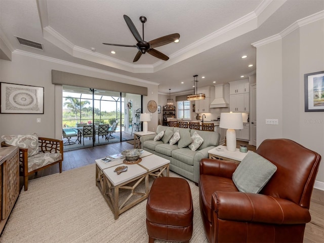 living area featuring crown molding, light wood-style flooring, visible vents, a tray ceiling, and a ceiling fan