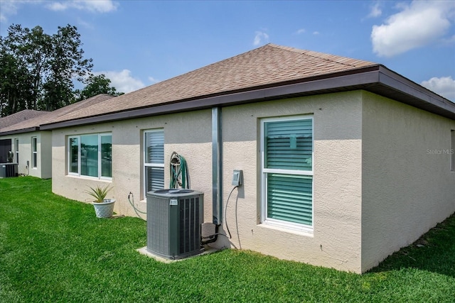 rear view of house with central AC unit, stucco siding, a yard, and a shingled roof