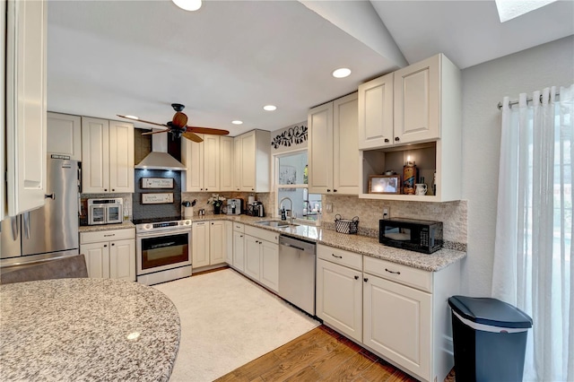 kitchen featuring a sink, stainless steel appliances, decorative backsplash, ceiling fan, and wall chimney range hood