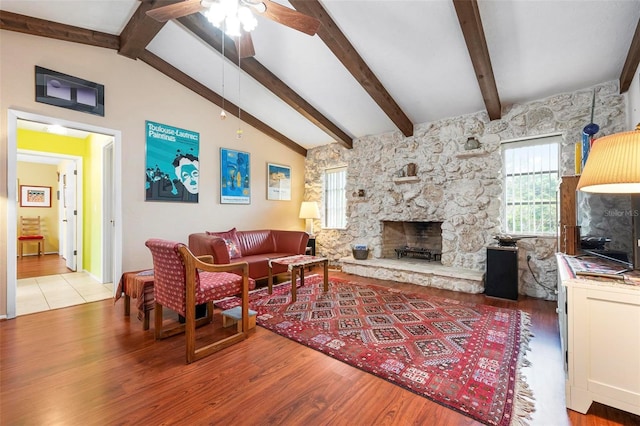living room with a ceiling fan, a stone fireplace, vaulted ceiling with beams, and wood finished floors