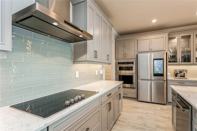 kitchen featuring stainless steel appliances, wall chimney range hood, decorative backsplash, light stone counters, and glass insert cabinets