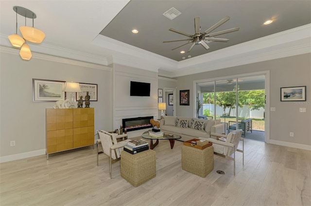 living room with a raised ceiling, a ceiling fan, light wood-type flooring, crown molding, and a fireplace