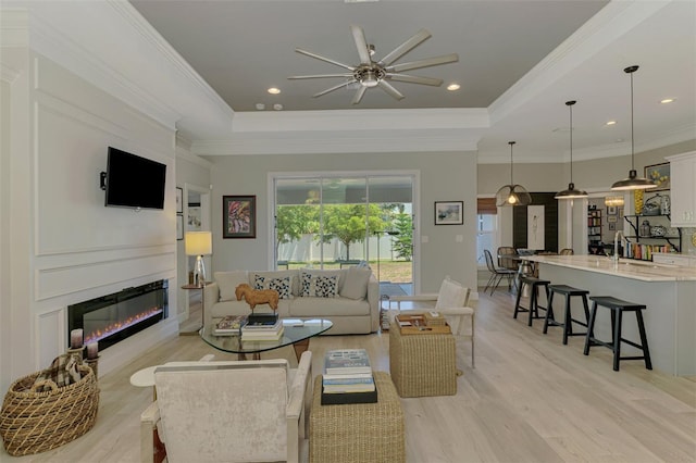 living room featuring a ceiling fan, light wood-style floors, a raised ceiling, a glass covered fireplace, and ornamental molding