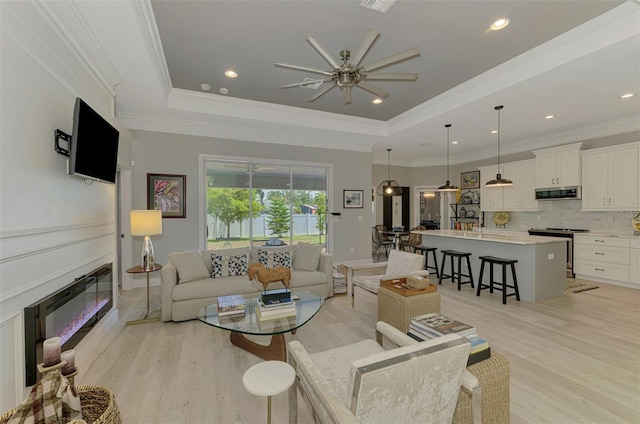 living area with a ceiling fan, light wood-style flooring, a raised ceiling, recessed lighting, and a glass covered fireplace