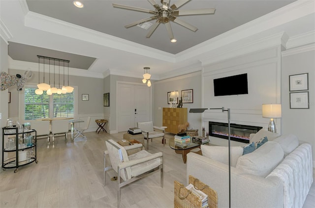 living room with a raised ceiling, a ceiling fan, light wood-style flooring, ornamental molding, and recessed lighting