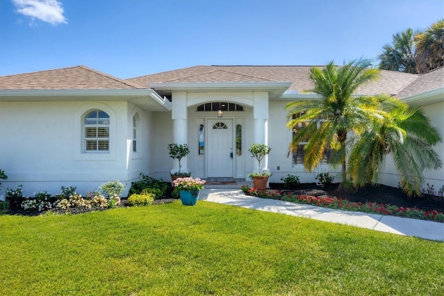 view of exterior entry with a lawn, roof with shingles, and stucco siding