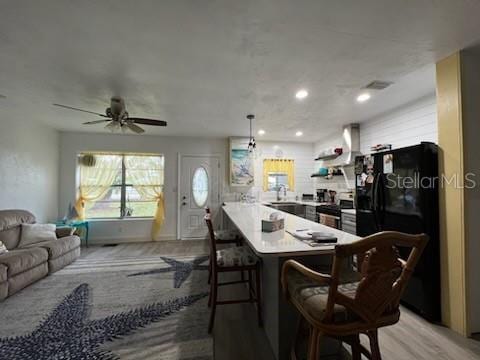 kitchen featuring black refrigerator, ceiling fan, wall chimney range hood, stainless steel range oven, and a breakfast bar