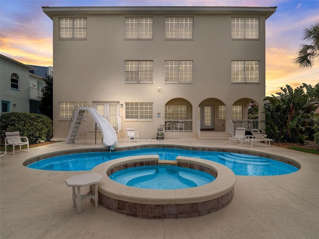pool at dusk featuring a water slide, a patio, and an outdoor hot tub