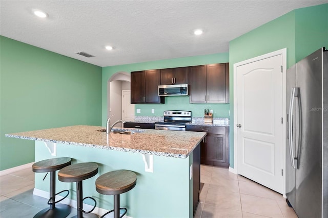 kitchen with sink, dark brown cabinets, a center island with sink, light tile patterned floors, and appliances with stainless steel finishes