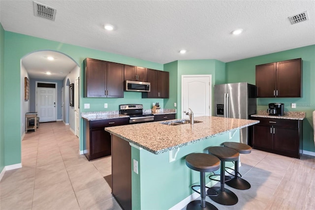 kitchen with dark brown cabinetry, sink, a center island with sink, light tile patterned floors, and appliances with stainless steel finishes