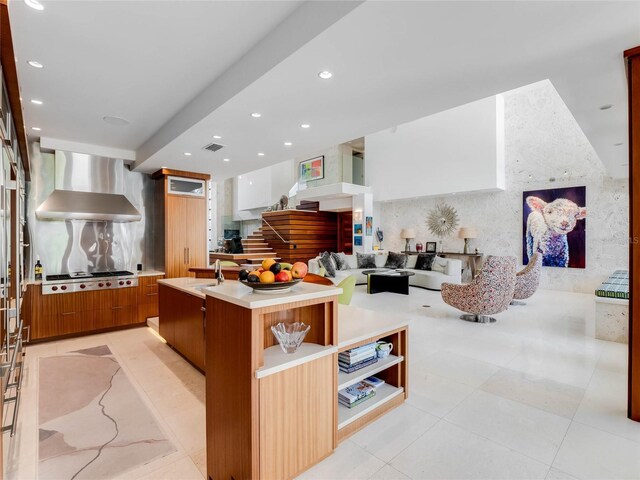 kitchen with sink, wall chimney exhaust hood, light tile patterned floors, an island with sink, and stainless steel gas cooktop