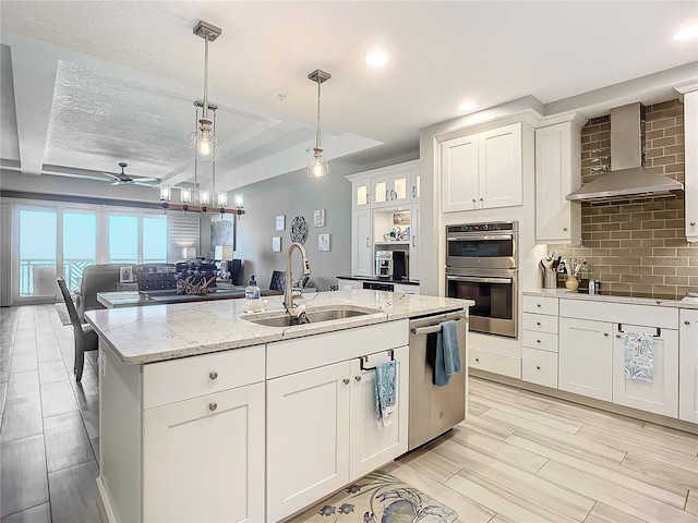 kitchen with a ceiling fan, a sink, backsplash, appliances with stainless steel finishes, and wall chimney range hood