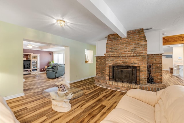 living room featuring beam ceiling, ceiling fan, a brick fireplace, light hardwood / wood-style floors, and a textured ceiling