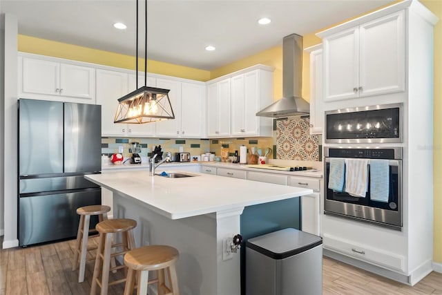 kitchen with a kitchen island with sink, sink, wall chimney range hood, decorative light fixtures, and stainless steel refrigerator