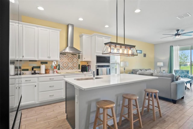 kitchen featuring white cabinetry, sink, wall chimney range hood, tasteful backsplash, and a center island with sink