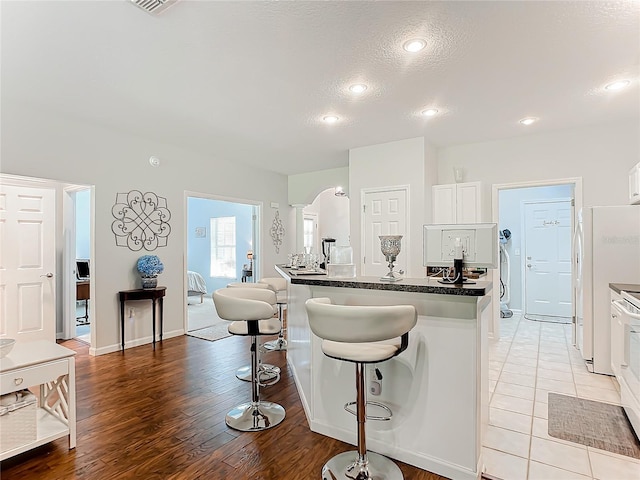 kitchen featuring a breakfast bar, arched walkways, dark countertops, white cabinets, and white appliances