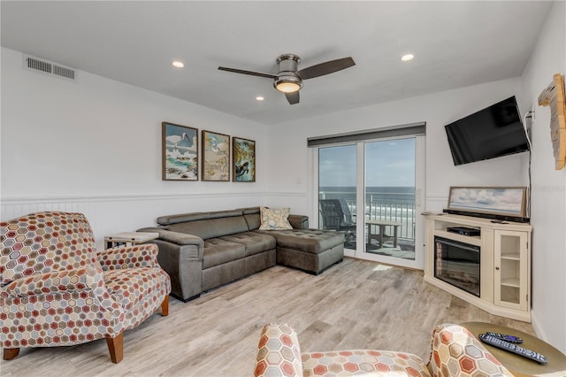 living area featuring a wainscoted wall, light wood finished floors, visible vents, a glass covered fireplace, and ceiling fan