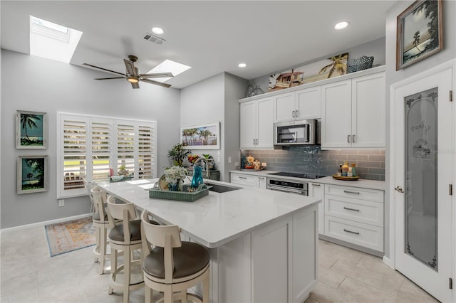 kitchen featuring appliances with stainless steel finishes, white cabinets, light stone countertops, a skylight, and a kitchen island with sink