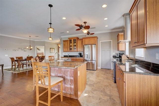 kitchen with hanging light fixtures, wall chimney exhaust hood, stainless steel appliances, ceiling fan, and dark stone counters