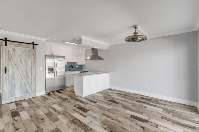 kitchen featuring white cabinets, stainless steel refrigerator with ice dispenser, a barn door, wall chimney exhaust hood, and crown molding