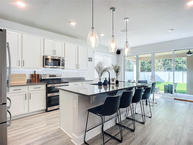 kitchen with pendant lighting, white cabinets, light wood-type flooring, an island with sink, and appliances with stainless steel finishes