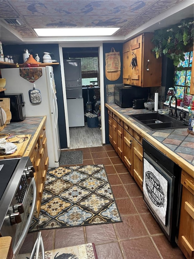 kitchen with black dishwasher, stainless steel stove, stacked washer / drying machine, tile counters, and sink