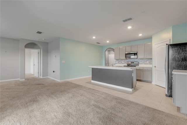 kitchen featuring a kitchen island with sink, gray cabinetry, light colored carpet, decorative backsplash, and appliances with stainless steel finishes