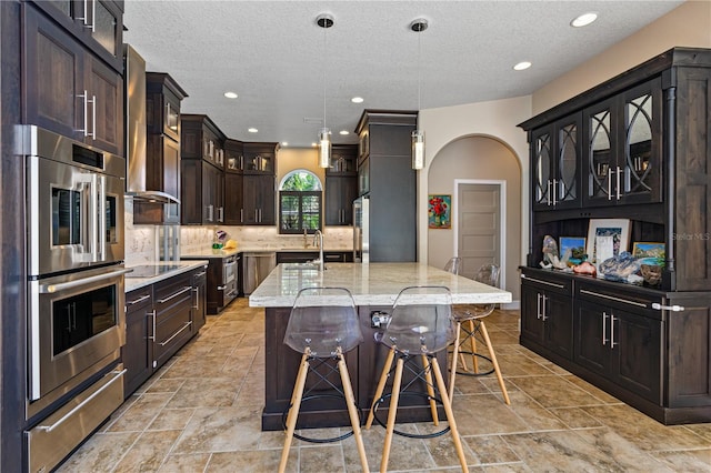 kitchen with a center island with sink, light stone counters, stainless steel double oven, and hanging light fixtures