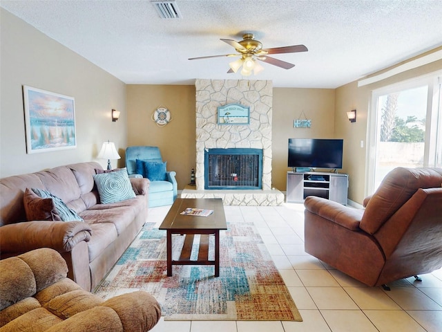 tiled living room with ceiling fan, a stone fireplace, and a textured ceiling